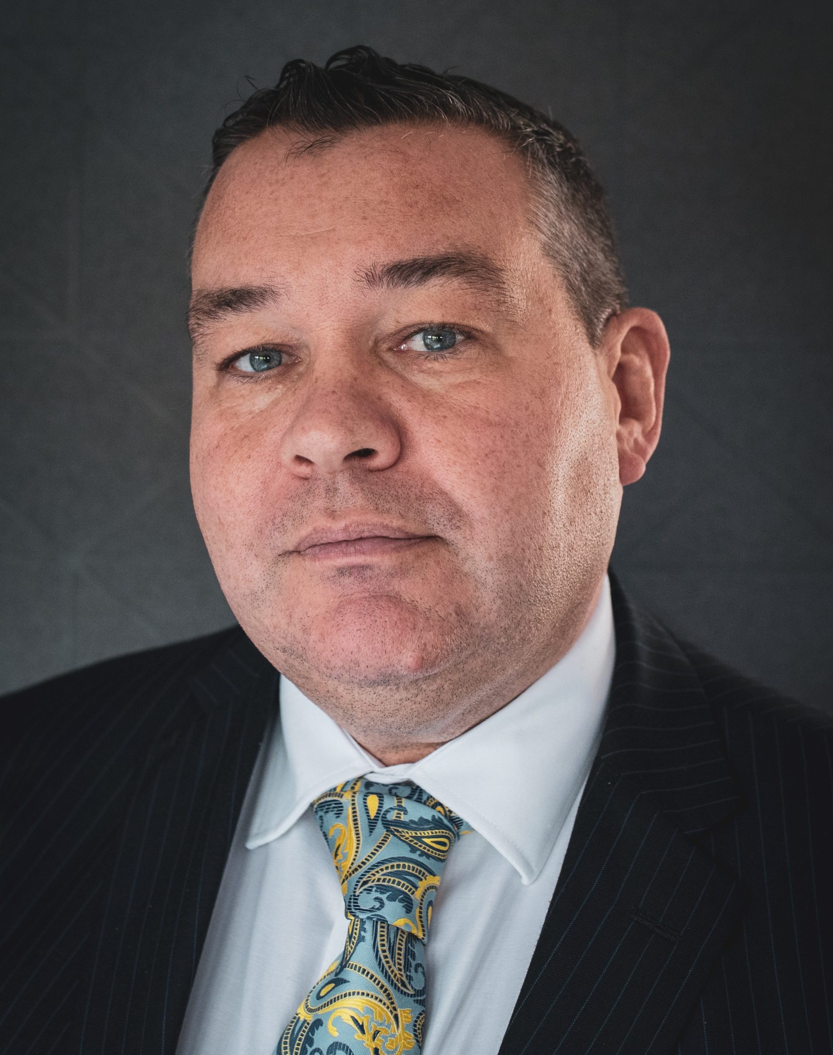 Close-up portrait of a man in a dark pinstripe suit and light blue patterned tie, looking slightly to the right against a dark backdrop.