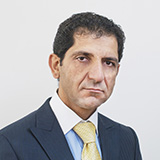 Professional portrait of a man in a navy suit and yellow tie against a light gray background, looking at the camera with a neutral expression.