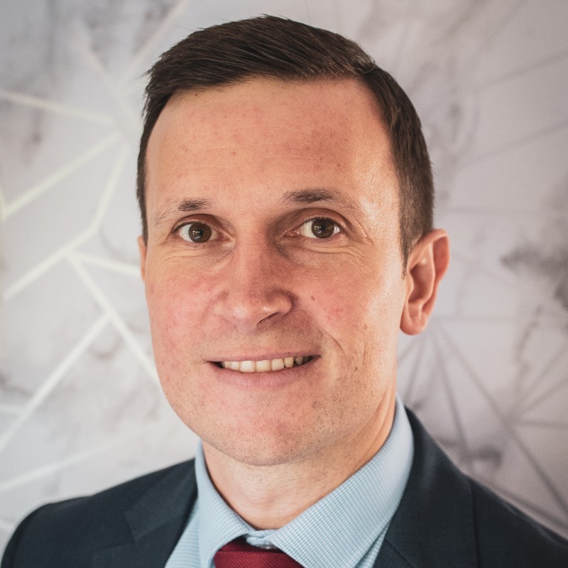 Richard Strike - Complyport - Your Trusted Partner in Governance, Risk, Compliance & Technology - Complyport - Your Trusted Partner in Governance, Risk, Compliance & Technology Portrait of a smiling man in a navy suit and light blue shirt, burgundy tie, against a light geometric backdrop.
