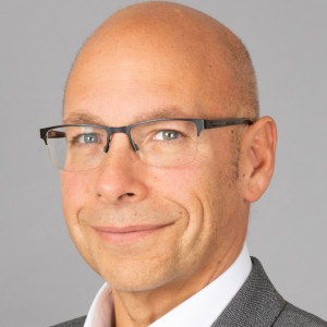 Close-up portrait of a smiling bald man wearing rectangular glasses and a gray suit against a gray background.