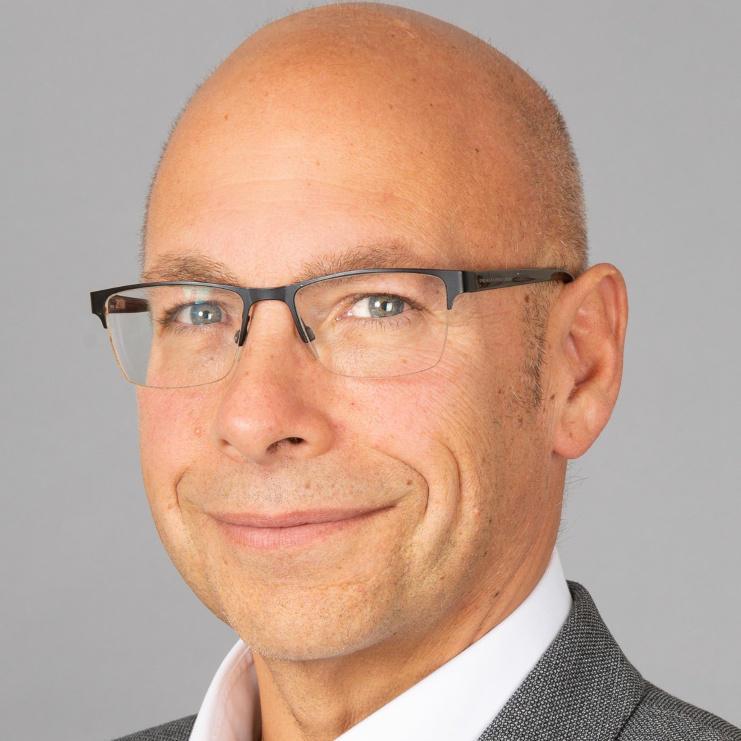 Close-up portrait of a smiling bald man wearing rectangular glasses and a gray suit against a gray background.
