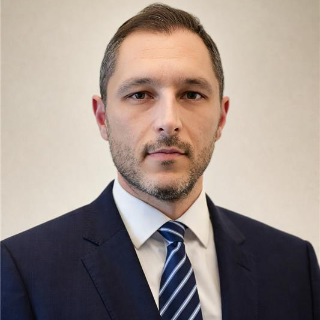 Professional headshot of a man in a dark suit, white shirt, and blue striped tie against a neutral background