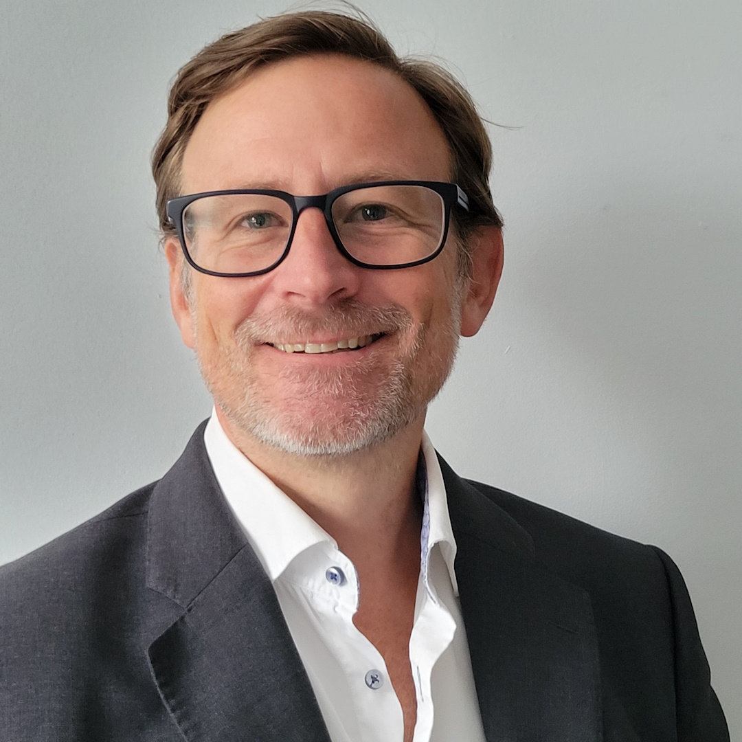 Smiling middle-aged man wearing glasses, a white shirt, and a dark blazer against a light-gray wall.