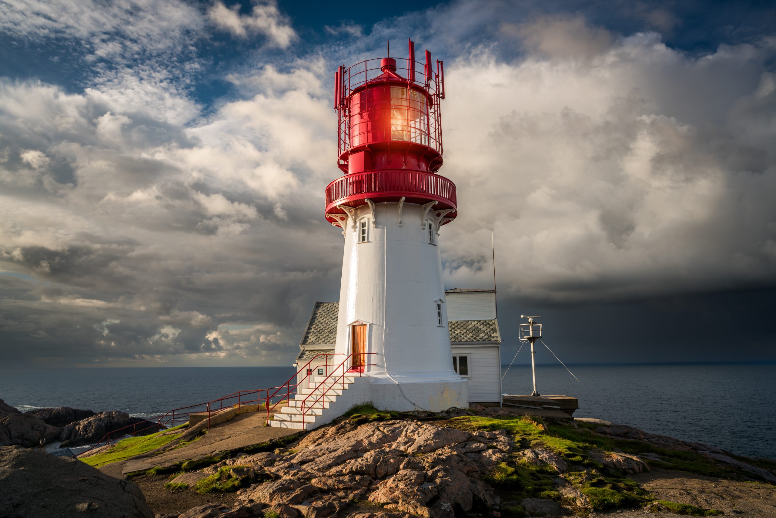 Red and white coastal lighthouse atop rocky shore with a stairway leading to an entry, overlooking dark sea and cloudy sky
