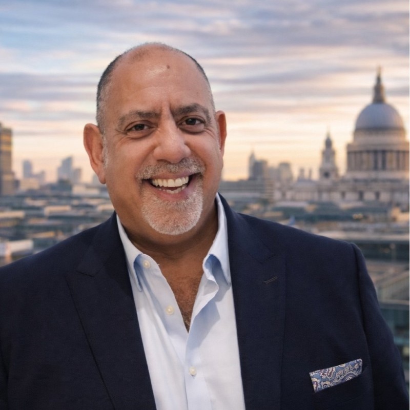 Smiling man in a navy blazer and light blue shirt, city skyline at dusk in the background, pocket square visible on his chest pocket.