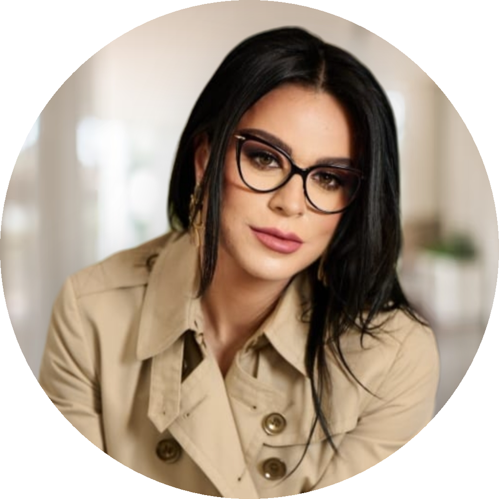 Portrait of a woman with dark hair and glasses, wearing a beige trench coat, looking at the camera in a softly blurred indoor setting