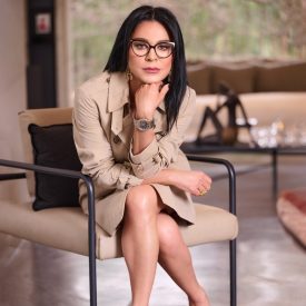 Professional woman with dark hair and glasses sits in a beige trench coat, hand under chin, in a modern lounge setting.